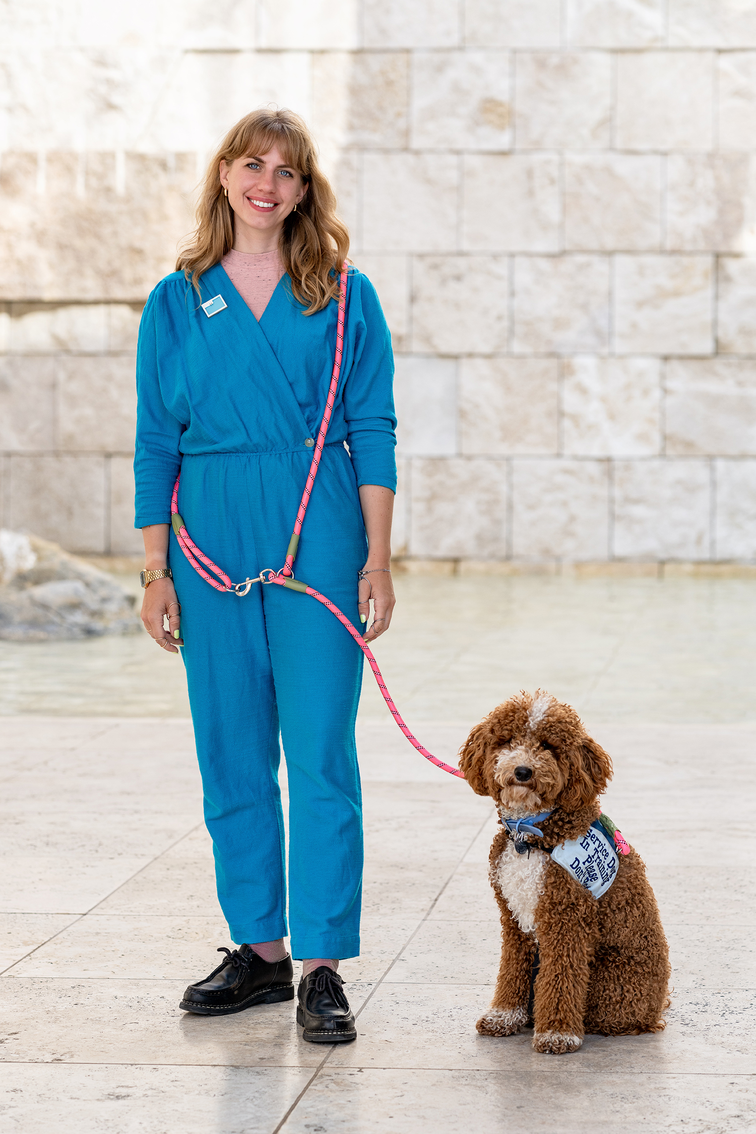 A thirty-something white woman with blond hair and bangs, wearing a teal jumpsuit and metal ring splints on her hands, standing against a tan travertine stone background. She has a bright pink leash around her torso, attached to a red-and-white miniature poodle wearing a denim service dog vest.